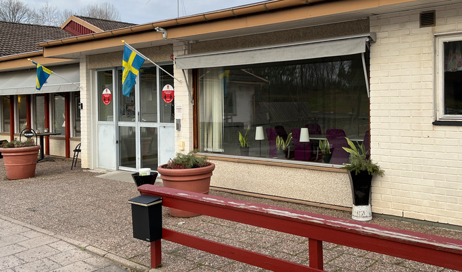 Entredörr vid Berggården i Rejmyre. På båda sidor om dörren hänger svenska flaggor. På höger sida finns ett rött staket med en brevlåda.